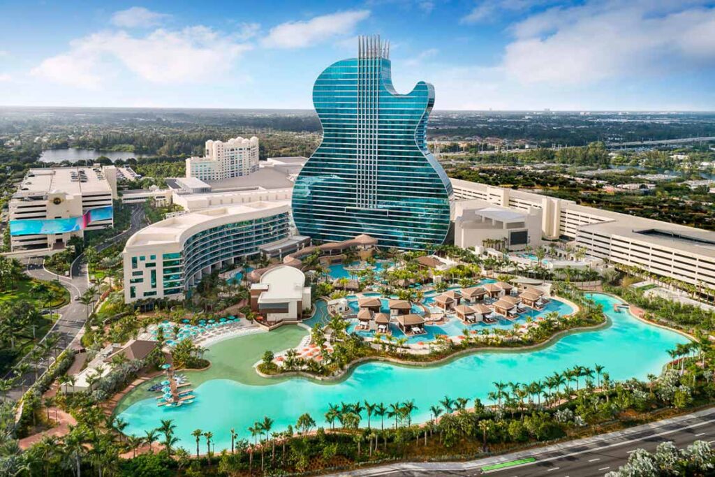 Aerial view of a large resort featuring a guitar-shaped building. The complex includes multiple structures, surrounded by lush palm trees. The centerpiece is a sprawling pool area with cabanas and water features, all set against an urban backdrop.