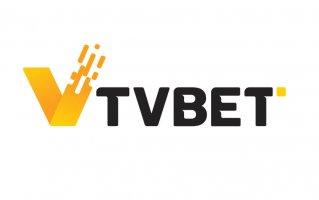 A imagem mostra o logotipo da TVBET. Ele apresenta uma letra "V" estilizada, amarela, com um design moderno e pixelado à esquerda, seguida pelo texto preto "TVBET" em letras maiúsculas e em negrito. O plano de fundo é branco.