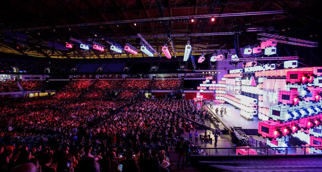 Uma arena grande e cheia acolhe um evento vibrante com um palco colorido e iluminado com vários ecrãs. Vários participantes enchem os assentos e um grupo de pessoas está de pé no palco. O teto da arena está decorado com luzes e vigas.